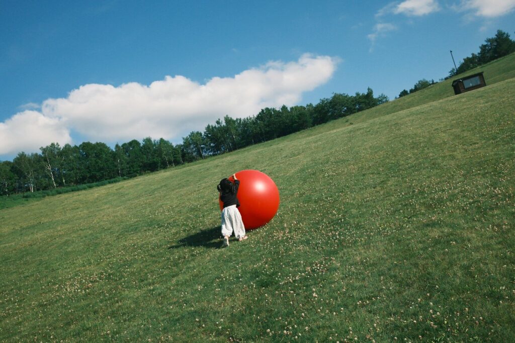 A child pushing a large red ball up a grassy hillside